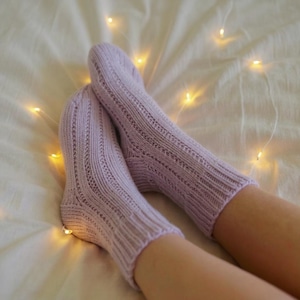 Puede incluir: Un par de calcetines de punto lavanda con detalles acanalados y calados. Los calcetines descansan sobre una superficie blanca, con luces de hadas cálidas en el fondo.
