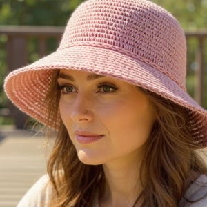 May include: A light pink, crocheted sun hat with a wide brim. The hat is made of a textured yarn and has a slightly rounded crown. The hat is designed to provide shade from the sun.