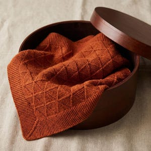 Op de afbeelding: Een open, ronde houten doos met een deksel, met een gevouwen, roestkleurige gebreide deken. De deken heeft een geometrisch ruitpatroon. De doos en deken staan tegen een neutrale achtergrond.