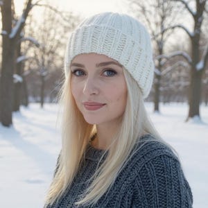 Könnte beinhalten: Eine Frau trägt eine weiße Strickmütze und einen grauen Pullover. Die Mütze ist gerippt und bedeckt den oberen Teil des Kopfes. Der Pullover hat ein strukturiertes Strickmuster. Der Hintergrund ist ein verschneiter Park mit kahlen Bäumen.