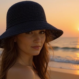 May include: A woman wearing a dark blue, wide-brimmed sun hat. The hat is made of a woven material. The background shows a beach at sunset, with the sun setting over the ocean. The sky is a warm orange color.