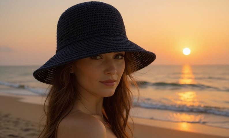 May include: A woman wearing a dark blue, wide-brimmed sun hat. The hat is made of a woven material. The background shows a beach at sunset, with the sun setting over the ocean. The sky is a warm orange color.