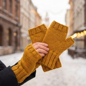 May include: Mustard yellow knitted fingerless gloves. The gloves are made of a thick yarn and have a ribbed cuff. The background shows a snowy street scene with buildings and string lights.