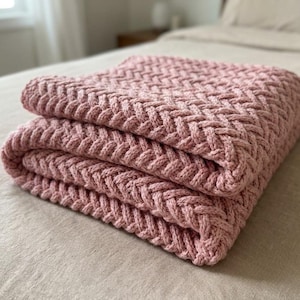 May include: A folded, dusky pink knitted blanket with a woven design. The blanket is made of a soft material and is folded neatly on a bed. The blanket is a cosy home decor item.
