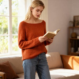 Może przedstawiać: Pomarańczowo-czerwony sweter z szerokim dekoltem i długimi rękawami. Sweter jest połączony z niebieskimi dżinsami. Osoba trzyma książkę. Otoczenie to dobrze oświetlony pokój z oknem i kanapą.