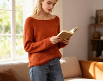 May include: An orange-red sweater with a wide neckline and long sleeves. The sweater is paired with blue jeans. The person is holding a book. The setting is a well-lit room with a window and a couch.