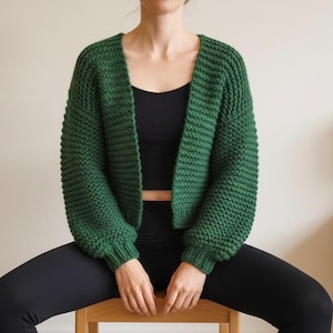Può includere: Un cardigan verde foresta a maglia grossa con maniche lunghe. Il maglione aperto è indossato sopra un top nero e abbinato a leggings neri. La trama del cardigan è visibile, mostrando il design a maglia.