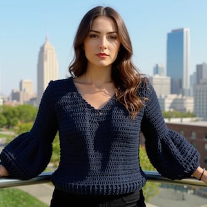 May include: A navy blue crochet jumper with a V-neck and long sleeves with flared cuffs. The jumper is made of a textured yarn and has a ribbed hem. The background features a cityscape with tall buildings.