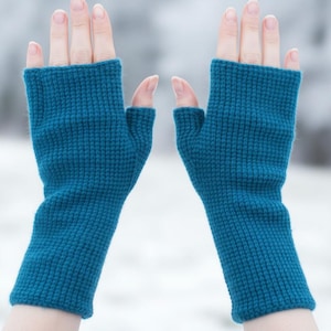 May include: A pair of teal fingerless gloves, knitted with a textured pattern. The gloves cover the hand up to the knuckles, leaving the fingers free. The background is a blurred, snowy landscape.