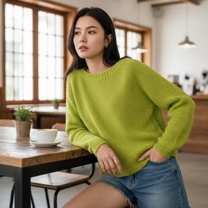 Könnte beinhalten: Eine Person trägt einen limettengrünen Pullover und Jeansshorts und sitzt in einem Café an einem Holztisch. Eine weiße Kaffeetasse und Untertasse stehen auf dem Tisch. Der Pullover hat einen Rundhalsausschnitt und lange Ärmel. Das Café hat große Fenster.