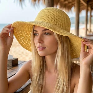 Puede incluir: Un sombrero de sol amarillo tejido con ala ancha. El sombrero es sostenido por las manos de una persona. El fondo muestra una playa con el océano y una mesa de madera.