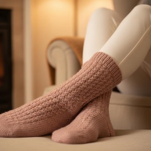 May include: A pair of knitted socks in a dusty rose colour. The socks have a textured pattern and are worn by someone sitting on a beige surface. The background includes a fireplace and a neutral-toned chair.