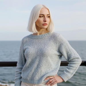 May include: A light blue sweater with a crew neck and long sleeves. The sweater has a textured knit design. The person in the image is wearing the sweater and is standing outside near the ocean.