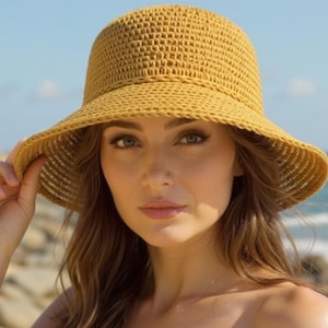 May include: A woman wearing a yellow crocheted sun hat with a wide brim. The hat is made of a woven material and has a rounded crown. The woman is outdoors, with a beach and ocean in the background.