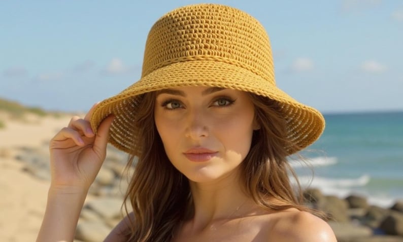 May include: A woman wearing a yellow crocheted sun hat with a wide brim. The hat is made of a woven material and has a rounded crown. The woman is outdoors, with a beach and ocean in the background.