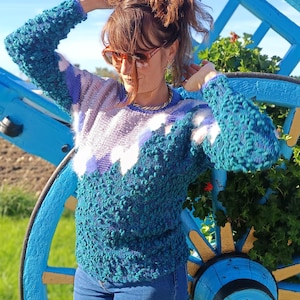 Peut inclure: Un pull texturé bleu sarcelle et violet avec un motif blanc et violet. Le pull a un col rond et des manches longues. La personne porte un jean bleu et des lunettes de soleil orange. Une roue de chariot bleue est en arrière-plan.