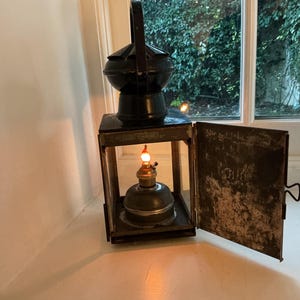 May include: A black metal lantern with a handle, a hinged door, and a lit oil lamp inside. The lantern is on a white surface near a window with greenery visible outside.