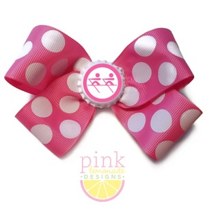 May include: A bright pink polka dot hair bow with white spots. The bow features a bottle cap center with a pink rowing team graphic. The text "pink lemonade designs" is at the bottom.