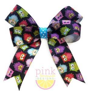 May include: A handmade hair bow featuring a black background with colorful owl illustrations. The bow has a light blue polka dot center and the text "pink lemonade designs" with a lemon graphic.