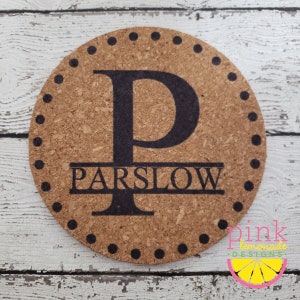 May include: Round cork coaster with a large black letter "P" and the name "PARSLOW" beneath it. The coaster has a dotted border and a light brown color. The bottom right corner has a logo that says "pink lemonade designs".
