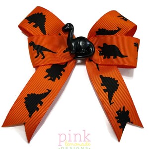 May include: Orange ribbon bow with black dinosaur silhouettes and a black dinosaur embellishment in the center. The bow is a handmade accessory, perfect for adding a touch of fun to any outfit. The bow is approximately 4 inches wide.