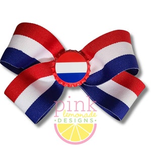 May include: A patriotic hair bow featuring red, white, and blue striped grosgrain ribbon. The center of the bow is a bottle cap with a red, white, and blue design. The bow is a decorative accessory.