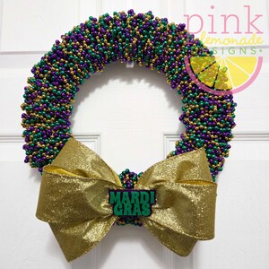 May include: A Mardi Gras wreath with a gold bow and a sign that says "Mardi Gras". The wreath is made of purple, green, and gold beads. The bow is made of gold glitter fabric.