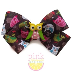 May include: A brown hair bow with a repeating owl pattern in pink, green, blue, and white. A yellow owl embellishment with green wings and a pink body is centered on the bow. The text "pink lemonade designs" is at the bottom.