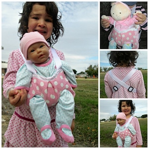 May include: A young girl wearing a pink and white polka dot dress carries a doll in a pink and white polka dot baby carrier. The doll is wearing a blue and white outfit and a pink hat.