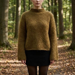 May include: A woman wearing a mustard-colored, long-sleeved sweater with a high neckline and a black skirt. The sweater has a fuzzy texture. The background is a forest with trees and fallen leaves.