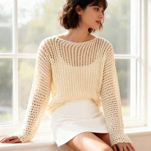 Op de afbeelding: Crèmekleurige trui met open weefsel, lange mouwen en een boothals. De trui wordt gedragen met een witte rok. Het breiwerk heeft een lichtgewicht, ademend ontwerp, geschikt voor warmer weer. Het model zit bij een raam.