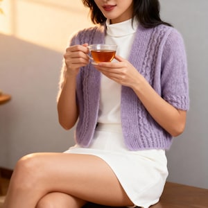 以下が含まれることがあります： ケーブル編みのデザインが施された、薄紫色の半袖カーディガン。白いタートルネックと白いスカートの上に着用されています。透明なガラスのティーカップが手に持たれています。