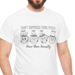 Puede incluir: Camiseta blanca con un gráfico de cuatro tazas de café de dibujos animados, cada una representando una emoción diferente: estrés, calma, cansancio y emoción. El texto "DON'T SUPPRESS YOUR FEELS" y "brew them honestly" están arriba y abajo de las tazas.