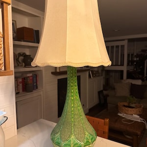 May include: A vintage table lamp with a green, textured ceramic base and a cream-colored lampshade. The base has a bulbous shape with a wooden base. The lampshade has a scalloped edge. The lamp is plugged into an electrical outlet.