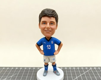 May include: A bobblehead figurine of a soccer player wearing a blue jersey with the number 13, white shorts, and blue socks. The figurine has a smiling face and is standing on a white circular base. The jersey has the Italian flag on it.