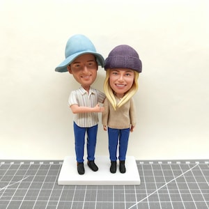 May include: A custom bobblehead figurine of a smiling couple. The man wears a light blue hat, striped shirt, and blue jeans. The woman wears a purple beanie, tan sweater, and blue jeans. Both figures stand on a white base.