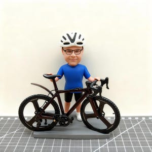 Könnte beinhalten: Figur einer Person mit weißem Fahrradhelm und Brille, die ein braunes Fahrrad hält. Das Fahrrad hat schwarze Reifen und einen schwarzen Sattel. Die Person trägt ein blaues T-Shirt und schwarze Shorts.