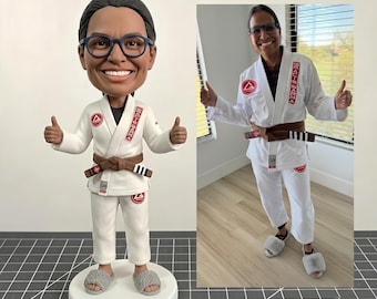 May include: A custom bobblehead figurine depicting a person wearing a white Brazilian Jiu-Jitsu gi with a brown belt, dark-rimmed glasses, and gray fuzzy slippers, standing on a round white base, next to a reference photo of the same person.