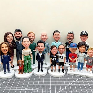 May include: A collection of custom bobblehead figurines, each depicting a person or couple. The figures are on white bases, with detailed facial features and clothing. Some figures are holding objects, and some have text on their bases.