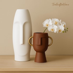 Puede incluir: Un jarrón blanco de cerámica con forma de rostro y un jarrón marrón de cerámica con asas doradas. El jarrón marrón contiene orquídeas blancas. Los jarrones tienen un diseño minimalista, con rasgos faciales. El logotipo de The Drifted Home está en la esquina superior derecha.