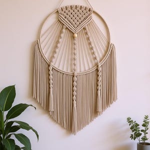 Op de afbeelding: Een beige macramé wandkleed met een cirkelvormig ontwerp. Het stuk heeft een geweven bovenstuk, een houten kraal en lange, verticale franjes. De macramé hangt aan een lus aan de bovenkant.