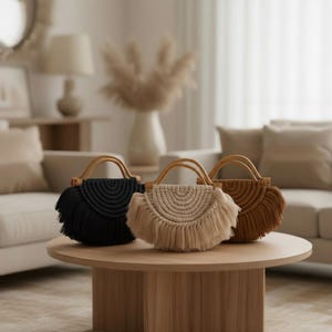 May include: Three macrame handbags in black, beige, and brown sit on a wooden table. Each bag has a half-circle design with fringe and a bamboo handle. The bags are a stylish accessory.