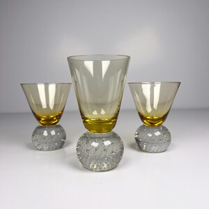 Puede incluir: Tres copas de cóctel vintage. Las copas tienen un tinte amarillo y se asientan sobre bases transparentes con textura de burbujas. Las copas de la izquierda y la derecha son del mismo tamaño, mientras que la copa central es más alta.