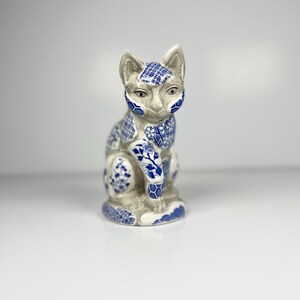 May include: A ceramic cat ornament in a seated pose. The cat is off-white with blue floral and geometric patterns on its face, ears, and body. The figurine has a detailed design and a glossy finish, suitable for home decor.