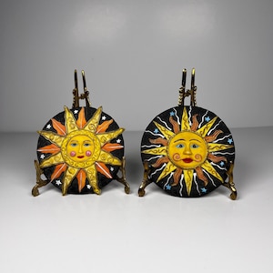 Peut inclure: Deux plaques circulaires représentant des visages de soleil jaunes avec des rayons orange et jaunes sur un fond noir parsemé de petites étoiles blanches et bleues. Chaque plaque est posée sur un chevalet en métal doré aux détails précis.
