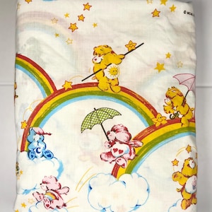 Puede incluir: Sábana blanca con personajes de Care Bears, arcoíris, nubes y estrellas. Los Care Bears están en varias poses, algunos sosteniendo paraguas. La sábana tiene un diseño colorido con acentos amarillos, rojos, azules y rosas.