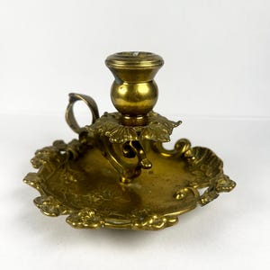 May include: A vintage brass candlestick holder with a round base and ornate floral detailing. The candlestick has a small handle and a cup to hold a candle. The brass has a warm, golden hue.