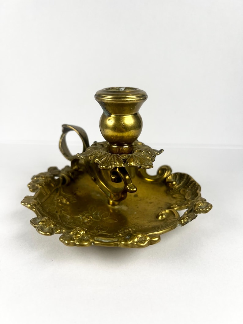 May include: A vintage brass candlestick holder with a round base and ornate floral detailing. The candlestick has a small handle and a cup to hold a candle. The brass has a warm, golden hue.