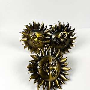 Puede incluir: Tres pomos decorativos con forma de cara de sol de color bronce, con una cara sonriente y rayos puntiagudos. Los pomos están dispuestos sobre una superficie blanca. Estos pomos son probablemente para muebles o fines decorativos.