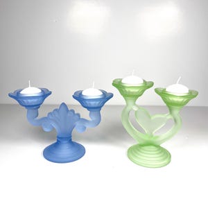 Peut inclure: Deux bougeoirs décoratifs. L'un est bleu avec un motif floral, contenant deux bougies. L'autre est vert, en forme de cœur, et contient deux bougies. Les deux sont en verre dépoli et ont des bougies blanches.